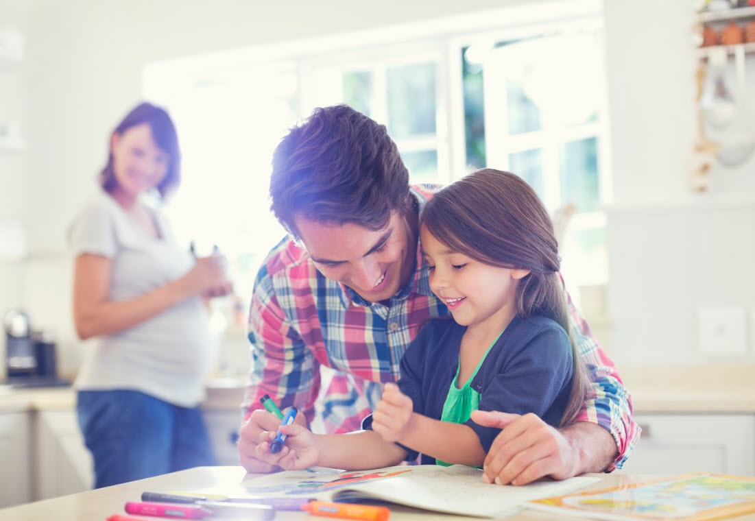 A father helps his daughter with colouring at a kitchen table while a pregnant mother smiles at the counter