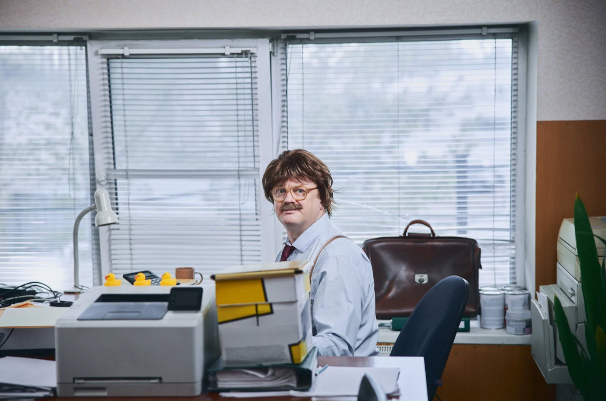 A man with a moustache and what appears to be a wig sits at an office desk