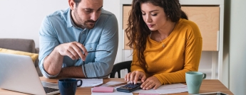 A couple calculates their bills over coffee at a kitchen table with a laptop and cacluclator