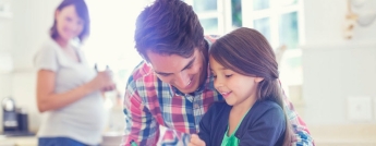 A father helps his daughter with colouring at a kitchen table while a pregnant mother smiles at the counter