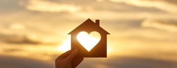 A person holds a cutout of a house with a heart-shape in the middle with the sun shining through