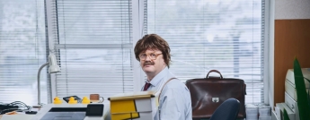 A man with a moustache and what appears to be a wig sits at an office desk